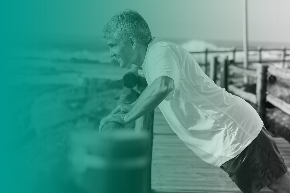 An man smiles an d supports men's health as he does a push up against an ocean boardwalk fence.