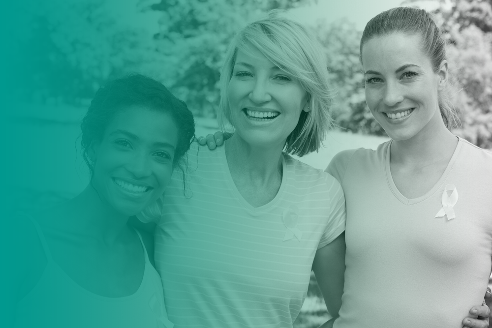 Three woman stand in a park with their arms around each other and smile. They wear breast cancer awareness ribbons to promote women's health.